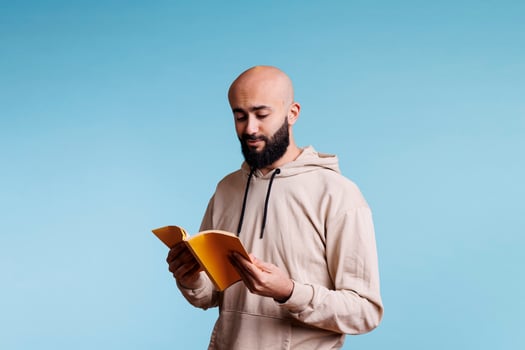 Arab man holding open book and reading novel with carefree expression. Young smiling bald bearded arabian person in hoodie standing with yellow softcover notepad and learning photo