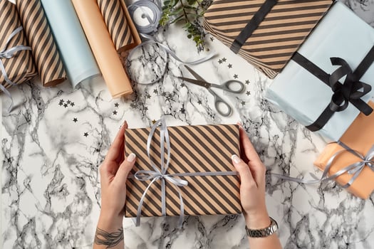 Hands of a woman are holding brown striped paper gift box tied with silver ribbon and bow against marble background. Close-up, copy space, top view. photo