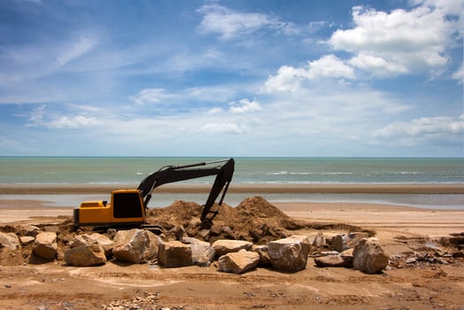 An excavator on a sandy beach, piling up dirt and rocks, with the ocean and a cloudy sky in the background. photo
