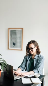 Young woman working on laptop in professional office setting. Student girl multitasking at home office. Work-from-home, freelance, business lifestyle concept. Technology, productivity and remote work. photo