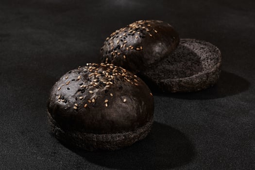 Fresh, baked, whole and cut in half black or chocolate bun sprinkled with sesame seeds against black background with copy space. Close-up photo