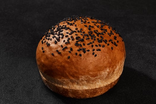 Fresh, palatable baked bun sprinkled with sesame seeds against black background with copy space. Rural cuisine or bakery. Close-up photo
