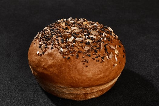 Fresh, palatable baked bun sprinkled with sesame and sunflower seeds against black background with copy space. Rural cuisine or bakery. Close-up photo