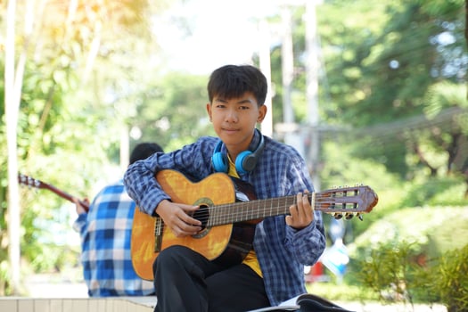 Asian boy happily sits outside playing guitar with his camp friends during break. Soft and selective focus. photo