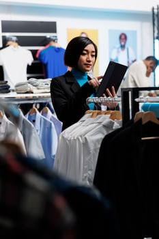 Clothing store asian woman employee using digital tablet while working in shopping center. Department mall boutique worker examining apparel in stock and managing inventory photo