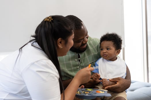 Asian mother feeding her 9 months old her cute little baby and African American helping for holding food plate At Home. Photo series of family, kids and happy people concept. Parents feed kids.