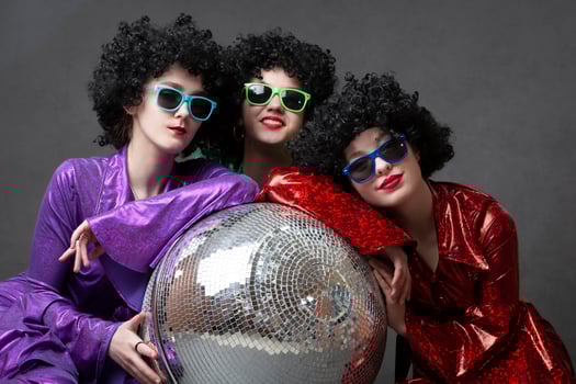 A group of disco girls in wigs with a disco ball and colorful costumes pose against a gray background. Disco party. photo