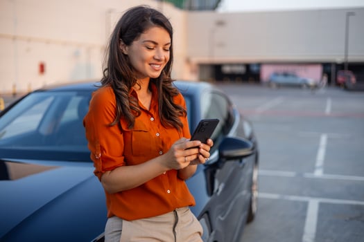 Successful smiling attractive woman in formal smart wear is using her smart phone while standing near modern car outdoors photo