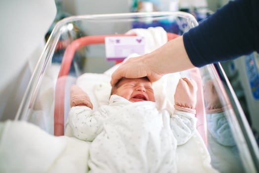 Newborn crying baby in hospital crib, father trying to calm down infant by putting his hand oh her head photo