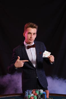 Elegant man is playing poker at casino. He is posing at the table with stacks of colored chips on it, against a black, smoke background. photo