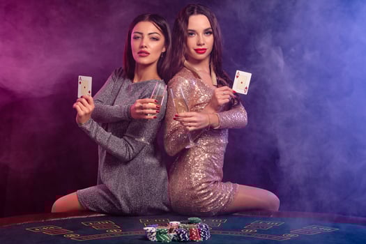 Two girls are playing poker at casino, holding glasses of champagne and cards, sitting on table with chips on it. Black, smoke background. Close-up. photo
