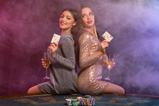 Two girls are playing poker at casino, holding glasses of champagne and cards, sitting on table with chips on it. Black, smoke background. Close-up. photo