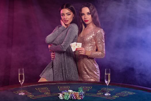 Two girls are playing poker at casino, hugging, holding cards, posing at a table with chips, champagne on it. Black, smoke background. Close-up. photo