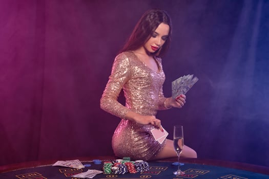 Girl playing poker at casino, holding money, cards. Sitting on table with chips, champagne, cash on it. Black, smoke background. Close-up. photo