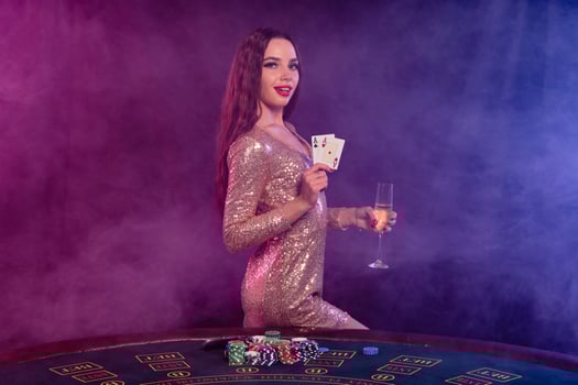 Girl playing poker at casino, holding glass of champagne, two aces. Posing at table with chips, money on it. Black, smoke background. Close-up. photo