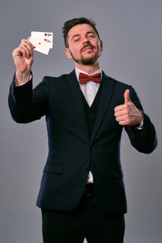 Man in black classic suit and red bow-tie showing two playing cards while posing against gray studio background. Gambling, poker, casino. Close-up. photo