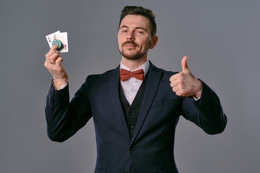 Man in black classic suit and red bow-tie showing two playing cards and chips, posing on gray studio background. Gambling, poker, casino. Close-up. photo