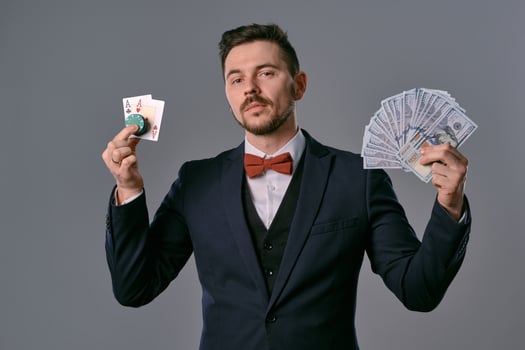 Man in black classic suit and red bow-tie showing two playing cards, chips and cash, posing on gray background. Gambling, poker, casino. Close-up. photo