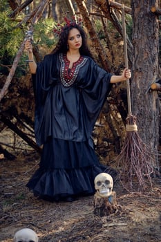 Witch in black, long dress, with red crown in her long hair. Posing with broom and skull in pine forest. Spells, magic and witchcraft. Full length. photo