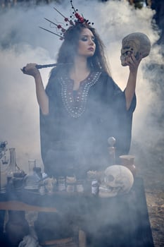 Witch in black, long dress, with red crown in her hair. Posing in pine forest, holding skull, making potion. Spells and witchcraft. Close-up, smoke. photo
