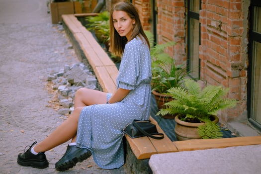 Blonde girl in long blue dress and small black handbag on her shoulder is posing sitting sideways on a parapet. Fashion and style. Full length shot. photo