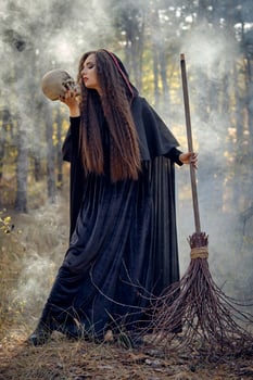 Witch in black, long dress with cape and hood. Posing in smoke pine forest. Holding a skull and a broom. Spells, magic and witchcraft. Full length. photo