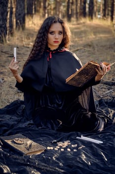 Witch in black dress with cape and hood. Posing in pine forest. Sitting on dark blanket whith books, candles and runes on it. Full length. photo
