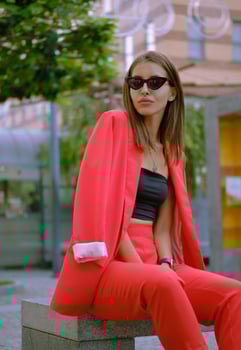 Blonde girl in a red lady-type pantsuit and black top, watch, ring, pendant and sunglasses is posing sitting on a stone bench. Close-up shot. photo