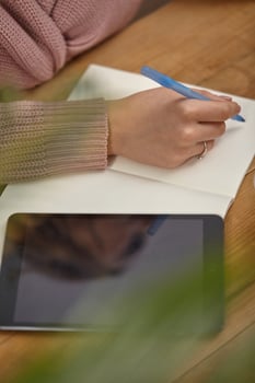 Model in pink cardigan. She is holding a blue pen, sitting at wooden table with a tablet, notebook and green flower on it. Student, blogger. Close-up photo