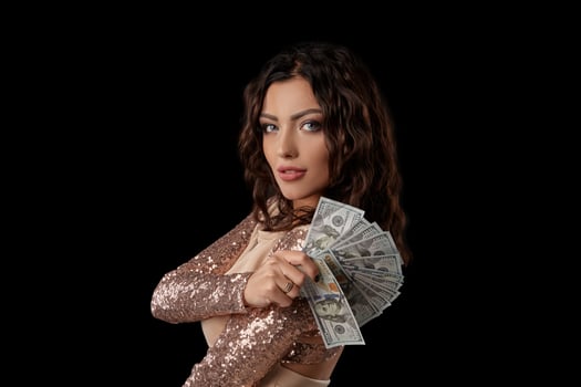 Brunette girl in shiny dress posing holding fan of hundred dollar bills in her hand standing on black studio background. Casino, poker. Close-up. photo