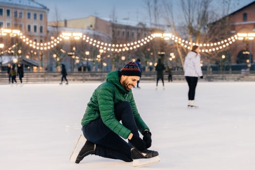 Man tying his ice skates on a festive lit rink at dusk, with skaters behind. photo