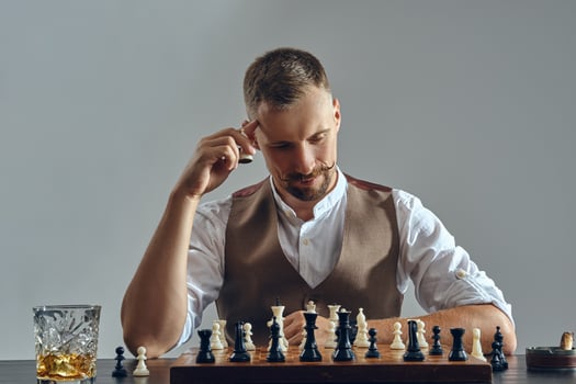 Man with stylish mustache, dressed in brown vest, white shirt is playing chess. Nearby glass of whiskey, ashtray, cigar. Grey background, close-up. photo