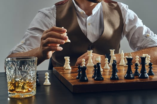 Hands of a man, dressed in brown vest, white shirt playing chess. Glass of whiskey is standing nearby. Grey background, close-up. photo