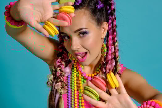 Lovely girl with multi-colored braids hairstyle and bright make-up, holding macarons between her fingers, posing in studio against a blue background. photo