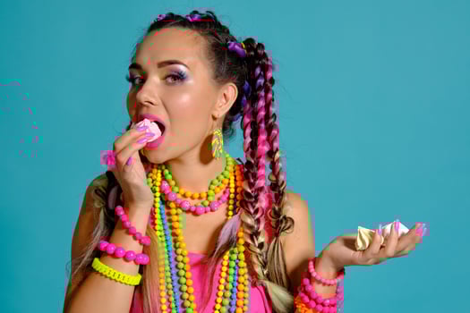 Lovely girl with a multi-colored braids hairstyle and bright make-up, posing in studio against a blue background, holding marshmallow in her hands. photo