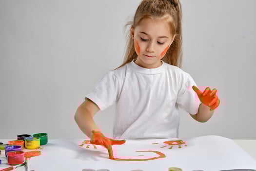 Little girl in white t-shirt sitting at table with whatman and colorful paints, painting on it with her hands. Isolated on white. Medium close-up. photo