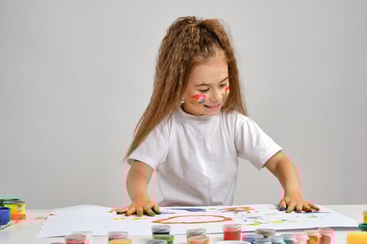 Little girl in white t-shirt sitting at table with whatman and paints on it, posing with painted face and hands. Isolated on white. Medium close-up. photo