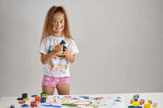 Little girl in white t-shirt standing at table with whatman and colorful paints, playing with a sponge soaked in paint. Isolated on white. Close-up. photo