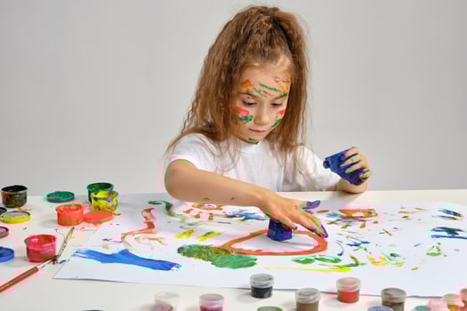 Little girl in white t-shirt sitting at table with whatman and paints, drawing on it, posing with painted face, hands. Isolated on white. Close-up. photo