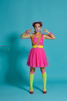 Lovely girl with a multi-colored braids hairstyle and bright make-up, posing in studio against a blue background, holding a lollipop in her hand. photo