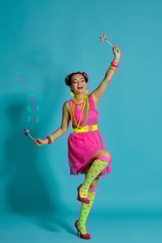 Lovely girl with a multi-colored braids hairstyle and bright make-up, posing in studio against a blue background, holding a lollipop in her hand. photo