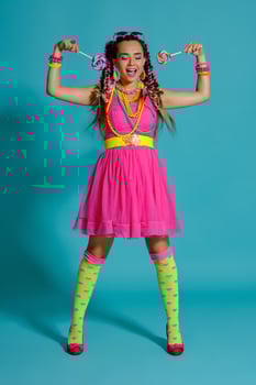 Lovely girl with a multi-colored braids hairstyle and bright make-up, posing in studio against a blue background, holding a lollipop in her hand. photo