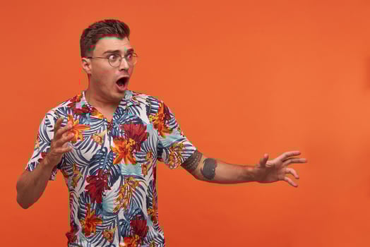 Young man in flowered shirt with wide opened mouth standing over the orange background, raising hands, looking away, wearing glasses, looks scared photo