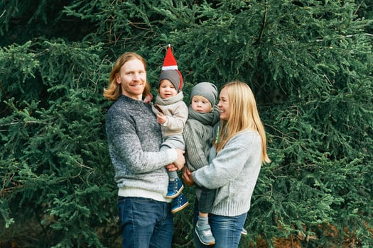 Outdoor portrait of beautiful happy young family of 4 posing in pine forest, wearing warm pullovers, couple with toddler boy and baby girl having good time at nature, cold weather, Christmas theme photo