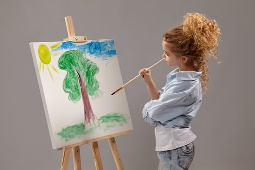 Charming school girl is painting with a watercolor brush on an easel, standing on a gray background. photo