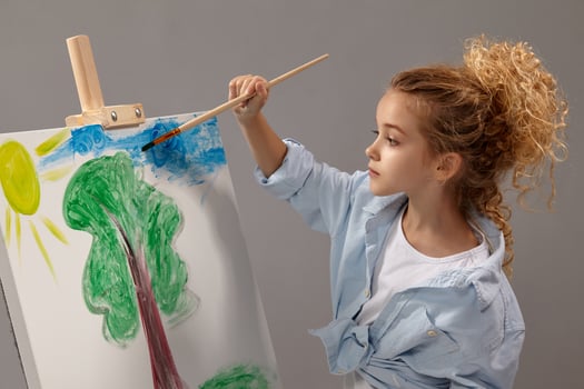 Charming school girl is painting with a watercolor brush on an easel, standing on a gray background. photo