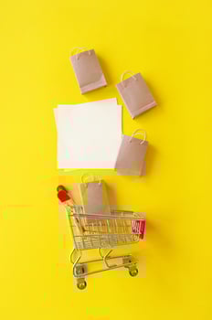 Mini shopping cart with small craft bags and blank white paper for lettering or shopping list on yellow background. photo