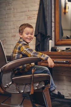 Portrait of a stylish little boy dressed in shirt and jeans in the barbershop, sitting in a chair against the barber's workplace photo