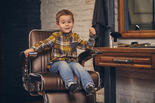 Portrait of a stylish little boy dressed in shirt and jeans in the barbershop, sitting in a chair against the barber's workplace photo