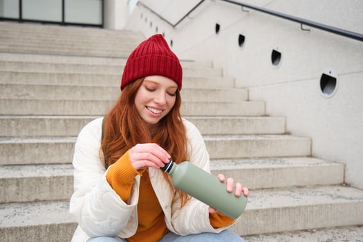 Smiling traveler, redhead girl tourist sits on stairs with flask, drinks hot coffee from thermos while travelling and sightseeing around foreign city, sits on stairs and rests photo